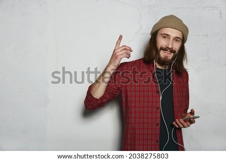 Young smiling european man listening music on headphones at mobile phone and having new idea. Stylish guy looking at camera. Isolated on white wall background. Studio shoot. Copy space