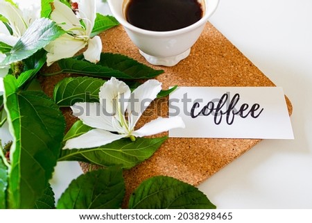 Black coffee cup on cork board, white flowers, card with the phrase: "coffee", and blackberry tree leaves on white background. Flat lay, Top view. Spring concept.