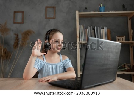 A positive little schoolgirl in headphones communicates with the tutor, waves her arms and greets the tutor during a video call via a laptop during an online lesson at home. Copy space.