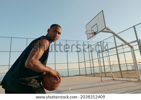 Young focused african sportsman holding basketball ball and looking away on sports court. Black man wearing sportswear. Urban basketball player. Sea behind mesh fence. Sunny daytime