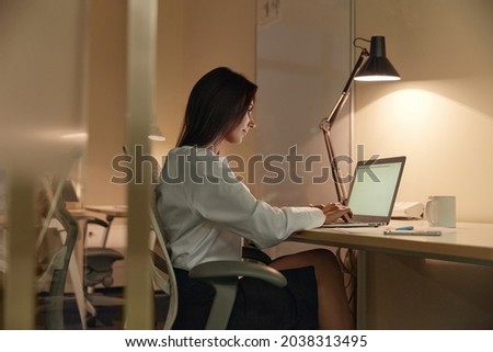 European businesswoman typing on laptop computer while sitting on chair at desk. Concept of overtime work. Young brunette woman wearing formal clothes. Office interior. Evening time Royalty-Free Stock Photo #2038313495