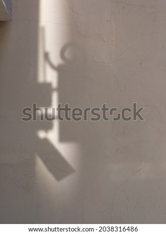 Roughly plastered with cracks, the wall of an old house with abstract shadows. Light brown textured grunge background with a vertical strip of sunlight. Light path, geometric shadows. Vertical photo.
