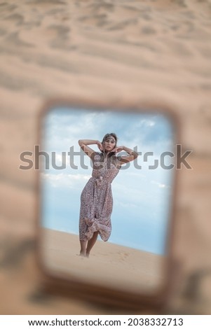 Photo in the mirror of attractive blond woman in long dress walking in desert. Wearing jewellery on her head. Picture in the mirror. Sand dunes. 