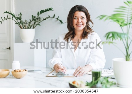 Portrait of Young smiling brunette woman doctor nutritionist plus size in white shirt working at laptop at modern bright office room Royalty-Free Stock Photo #2038354547