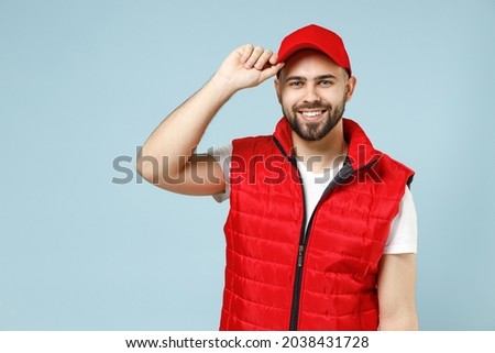 Professional happy delivery guy employee man in white T-shirt vest uniform workwear work as dealer courier hold touch red cap isolated on pastel blue color background studio portrait. Service concept