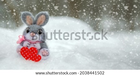 Happy Valentine's Day greeting card or banner. Plush rabbit holding a red heart ka symbol of love. Holidays Happy Valentine's Day. Snowfall on the background of the forest