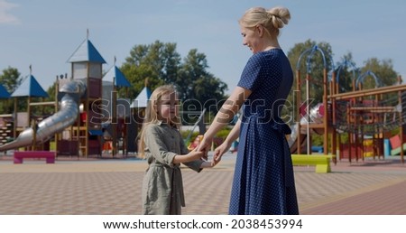 Side view of young mother and little daughter with long hair walking together holding hands in summer park. Happy caucasian mom and preschool child play at playground