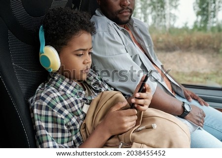 Serious Black curly-haired boy in bright headphones watching cartoon on phone while riding bus with father