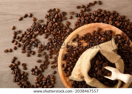 Sack full of coffee beans with kitchen shovel and pile of beans around on wooden table. Top view. Horizontal composition.