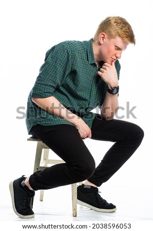 portrait of a red-haired guy in a green shirt and jeans on a white isolated background. teenager sitting on an old stool
