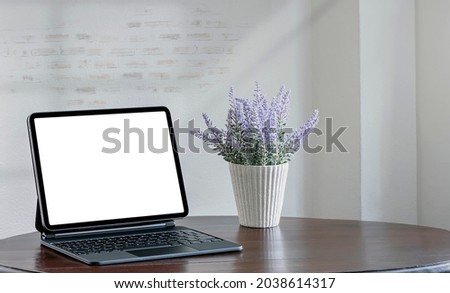 Blank screen tablet with magic keyboard and flower on wooden table in empty room.