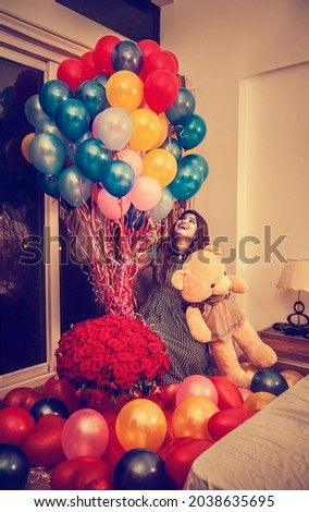 young girl sitting with bunch of red roses flowers, lots of colorful balloons, big teddy bear. happy women smiling face indoor. many balloons on the ground. birthday celebration. ready for party
