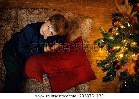 A blond boy 5-7 years old lies on the carpet near the New Year tree, looking at the camera. Top view. Selective soft focus, film grain effect