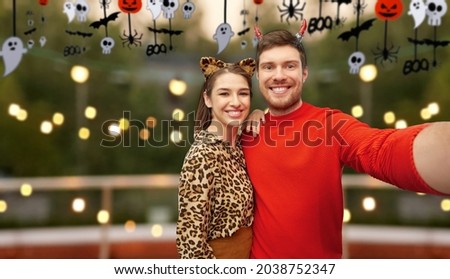 holiday and people concept - happy smiling couple in halloween costumes of devil and leopard taking selfie over roof top party background