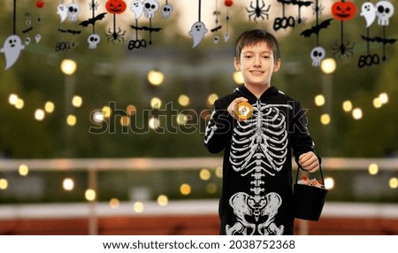 halloween, holiday and trick-or-treating concept - smiling boy in black costume of skeleton with candies and flashlight over roof top party background