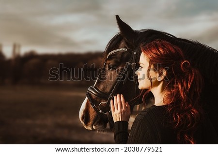 2 heads one love! stallion with head collar and redhead woman are bonding while being intimate friends and look left into sun to the future of cooperation of horse and rider enjoy the moment of trust Royalty-Free Stock Photo #2038779521