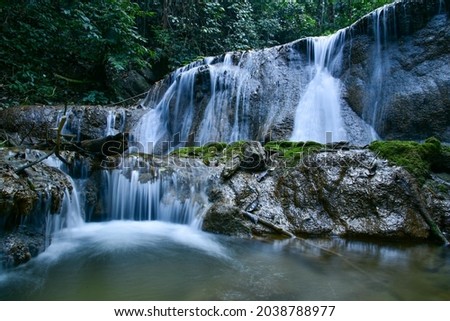 The waterfalls in the forest flowed in a soft white color, contrasting with the huge rocks of great power. Nearby there are big trees, light green, dark green, very natural.