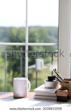 White classical ceramic mug mock up stock photography. Styled image of home office interior with large window and nature background. Writing supplies, large  and books on the desk.