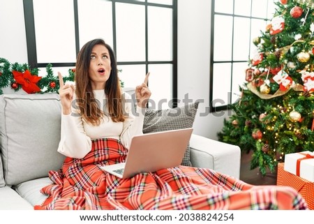 Beautiful caucasian woman using laptop sitting on the sofa by christmas tree amazed and surprised looking up and pointing with fingers and raised arms. 