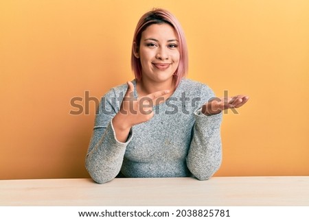 Hispanic woman with pink hair wearing casual clothes sitting on the table amazed and smiling to the camera while presenting with hand and pointing with finger. 