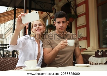 Young woman trying to take selfie with her displeased boyfriend in outdoor cafe. Boring date Royalty-Free Stock Photo #2038826000