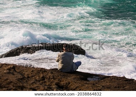 In the depth of field an old man sitting on a natural rock texture takes pictures of the wavy sea.