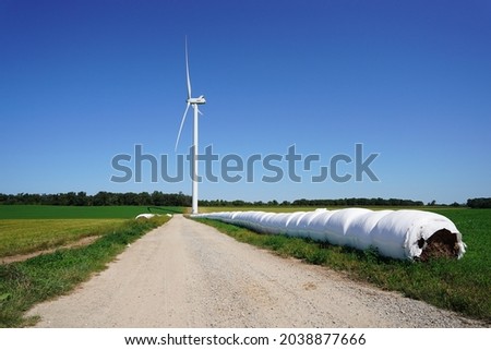 Wrapped hay bales align along dirt road in front of a giant wind turbine