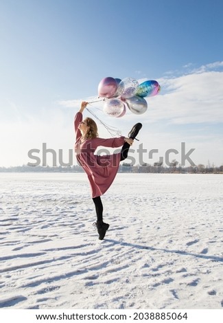 Full size photo of graceful teenage girl holding many colorful balloons in hands. Stands on one leg, raising other, in snow in pink dress. sunny winter day. Feels great, festive mood, enjoys moment.