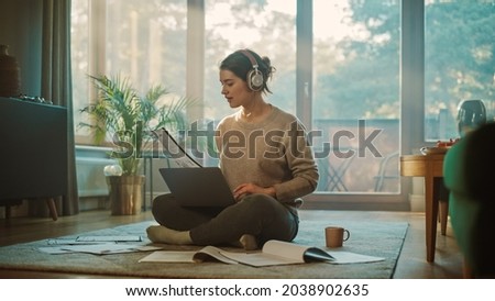 Young Woman Using Laptop at Home, Does Remote Work, Listens Music through Headphones. Beautiful Smiling Girl Sitting on the Floor Does Research on Papers, Documents, Brainstorms Creative Project Royalty-Free Stock Photo #2038902635