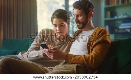 Couple Use Smartphone Device, while Sitting on a Couch in the Cozy Apartment. Boyfriend and Girlfriend Shopping on Internet, Watching Funny Videos, Use Social Media, Streaming Service. Royalty-Free Stock Photo #2038902734