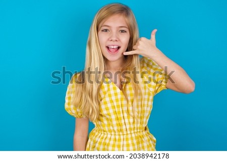 beautiful caucasian little girl wearing yellow dress over blue background makes phone gesture, says call me back again, has glad expression.