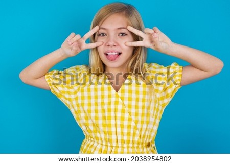 Cheerful positive beautiful caucasian little girl wearing yellow dress over blue background shows v-sign near eyes open mouth