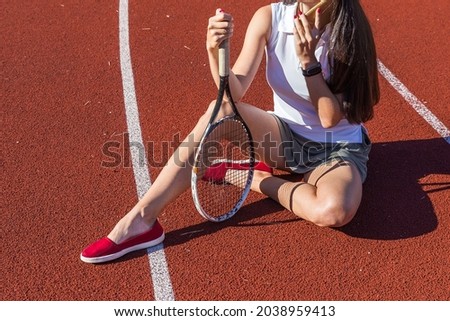 A model with a tennis racket sits on the court on a sunny day.