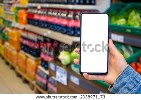 Woman hand holding mobile phone with blank white desktop screen while shopping at supermarket. Woman using smartphone, blurry supermarket in the background. Mock up image with copy space.