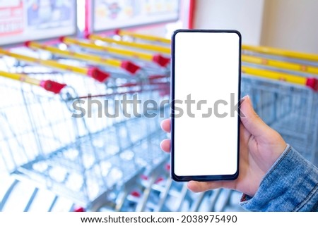 Mockup image of woman hand holding white desktop screen mobile phone in the supermarket. Many blurry shopping cart or trolley in the background.