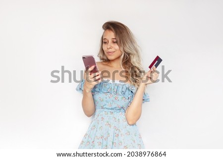 A young charming blonde woman with wavy hair in a blue dress holds a plastic credit card in her hand looking at the mobile phone screen while making an online purchase isolated on a white background