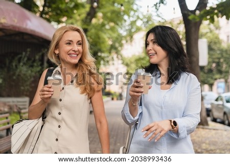 Two confident mature caucasian girlfriends women best friends sisters walking together and drinking hot beverage tea coffee in the morning outdoors in city park. Royalty-Free Stock Photo #2038993436