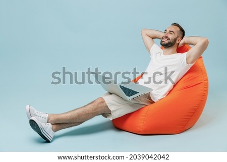 Full length young smiling happy man 20s in white t-shirt sit in bag chair hold use work on laptop pc computer hold hands behind neck rest relax isolated on plain pastel light blue background studio. Royalty-Free Stock Photo #2039042042