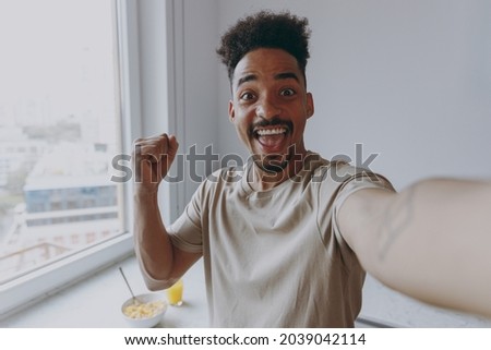Close up young african american man in casual clothes do winner gesture clench fist do selfie shot pov on mobile phone prepare cooking food in light kitchen at home alone indoor Healthy diet concept.
