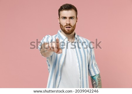 Young serious strict sad caucasian unshaven man 20s in blue striped shirt white t-shirt point index finger camera on you command do it isolated on pastel pink background studio. Tattoo translate fun.