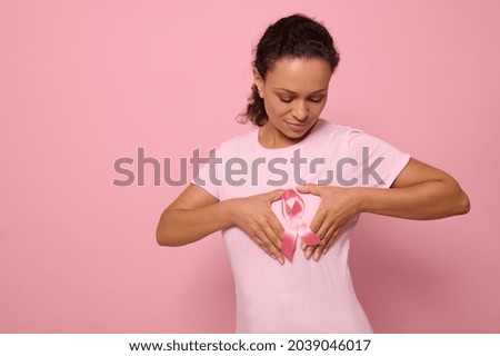 Isolated portrait on colored background of mixed race woman in pink t-shirt, putting her hands on her chest in shape of heart with a pink satin ribbon in the center. World Cancer Awareness Day. Royalty-Free Stock Photo #2039046017