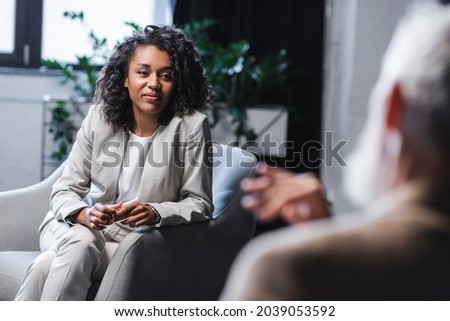 pretty african american journalist looking at blurred businessman sitting in armchair and gesturing while talking during interview Royalty-Free Stock Photo #2039053592