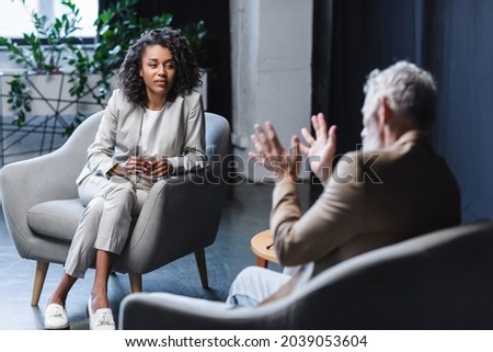 curly african american journalist looking at blurred businessman sitting in armchair and gesturing while talking during interview Royalty-Free Stock Photo #2039053604