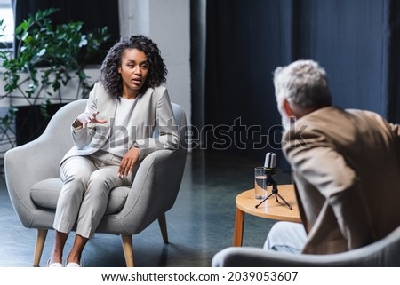 african american journalist gesturing while talking with blurred businessman sitting in armchair during interview Royalty-Free Stock Photo #2039053607