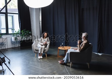 positive african american journalist in suit talking with businessman sitting in armchair during interview Royalty-Free Stock Photo #2039053616