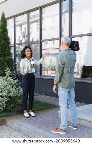 full length of cheerful african american journalist pointing with hand at building while doing reportage near cameraman Royalty-Free Stock Photo #2039053640