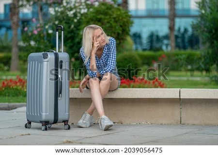 Young blonde woman in shorts, a shirt is sitting with a suitcase on a bench and thoughtfully looks away against the background of trees, bushes on a summer day