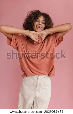 adorable dark-skinned girl posing on light pink background raising her hands to her face. curly-haired brunette with gentle smile is dressed in coral-colored T-shirt tucked into white jeans.