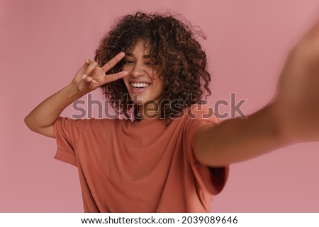 cute radiant lady makes selfie showing two fingers near her face on pink background. dark-haired, curly, smiling African woman with snow-white smile blinks one eye. Royalty-Free Stock Photo #2039089646