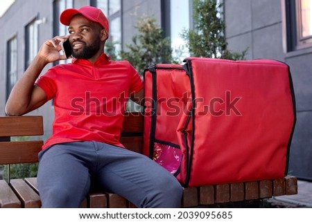 Black delivery man calling customer by smartphone outdoors. courier carrying food and drinks order on city street. Male in red uniform waiting for clients, sitting on bench alone, with thermo bag